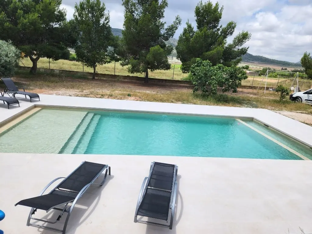 Piscina de agua clara con playa sumergida y escalones, con dos tumbonas negras sobre pavimento claro